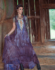 A woman wearing a long purple bohemian-style dress with fringe and a patterned bodice, standing inside a rustic wooden barn with hay bales and straw on the floor.